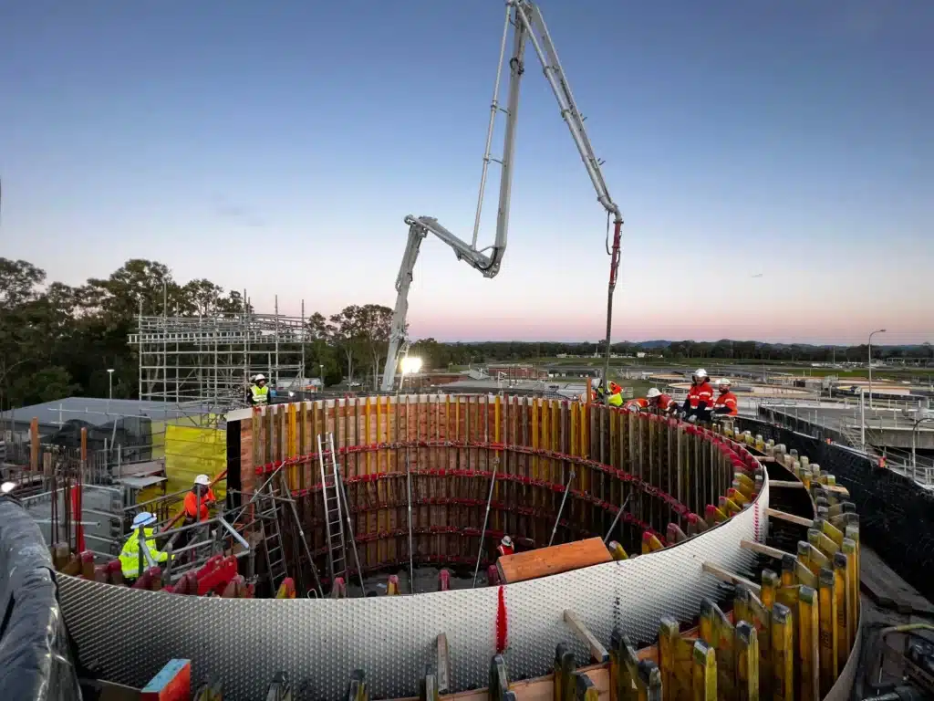 Construction workers operate machinery to pour concrete into a large circular foundation at a building site, surrounded by scaffolding and equipment, during sunset.