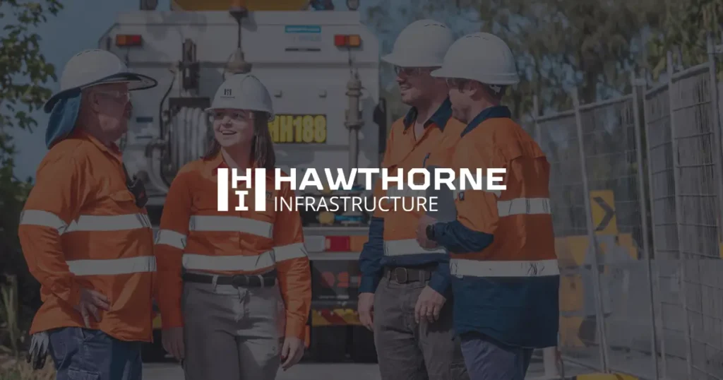 Four construction workers wearing hard hats and orange safety jackets stand and talk at a work site. The words Hawthorne Infrastructure appear over the image.