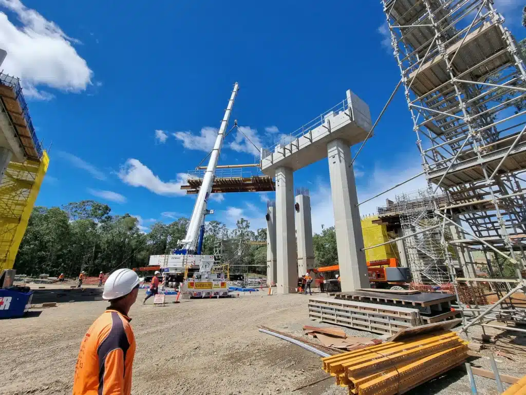 A construction site with scaffolding, large concrete pillars, a crane lifting materials, and workers in safety gear under a bright blue sky with scattered clouds.