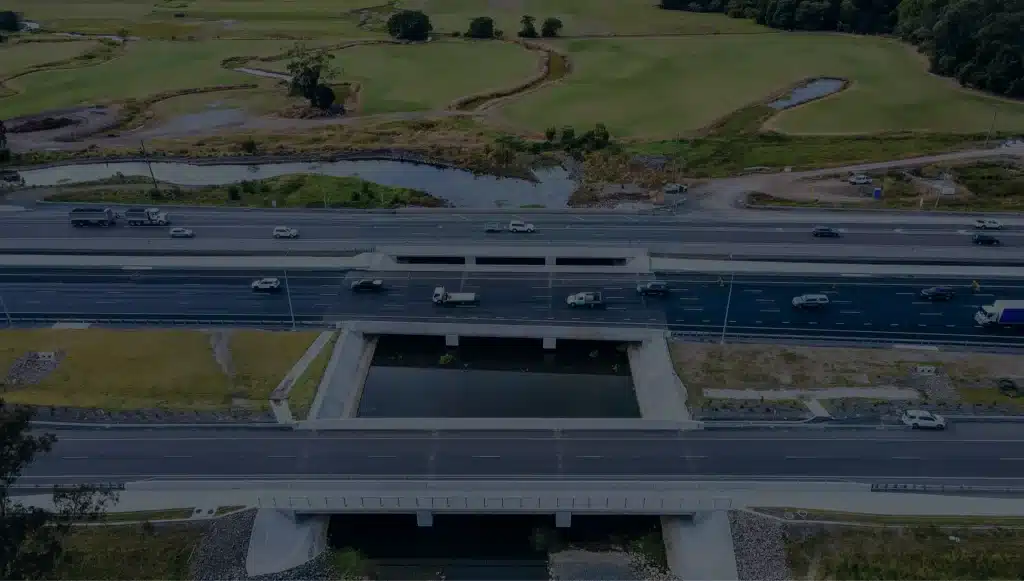Aerial view of a multi-lane motorway with cars and lorries, intersecting over a waterway and surrounded by green fields and trees.