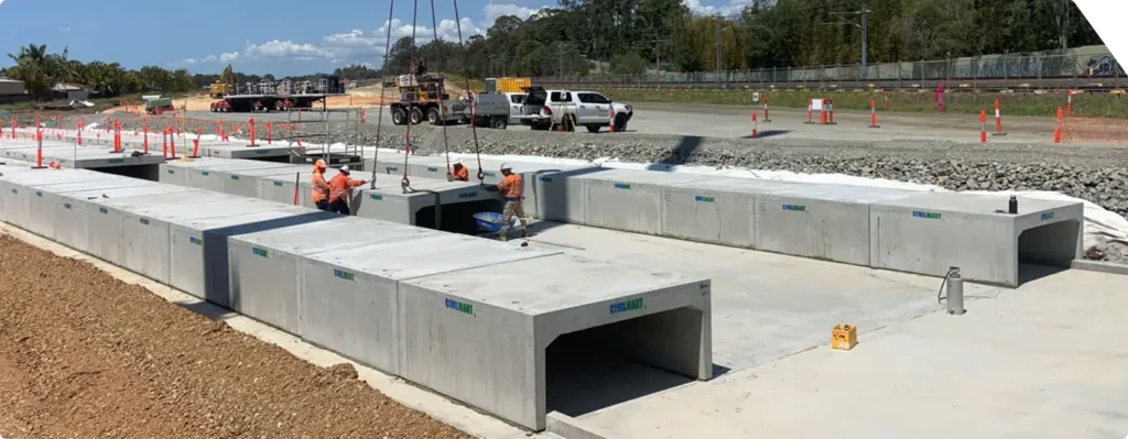 Construction workers in safety gear install large rectangular concrete culverts at a roadworks site, with lorries, orange cones, and machinery visible in the background under a partly cloudy sky.