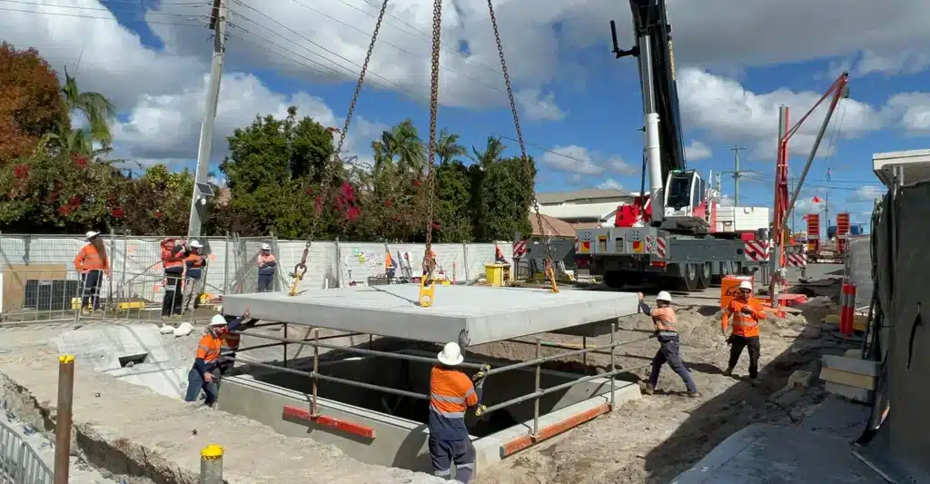 Construction workers use a crane to lower a large concrete slab into place at a work site. Several workers in safety gear guide the slab while others observe. Machinery and fencing are visible in the background.
