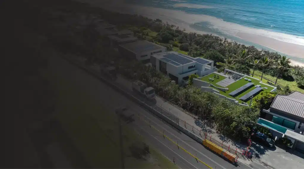 Aerial view of a coastal residential area with modern houses, green lawns, and a nearby road under construction, adjacent to a sandy beach and blue sea.