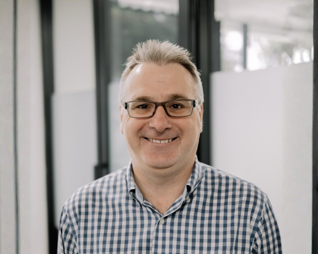 A middle-aged man with short grey hair and glasses smiles at the camera. He is wearing a blue and white checked shirt and standing indoors with glass panels in the background.