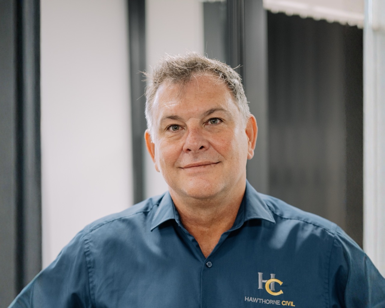 Middle-aged man with short grey hair wearing a blue shirt embroidered with HC Hawthorne Civil, standing indoors in front of a blurred office background.