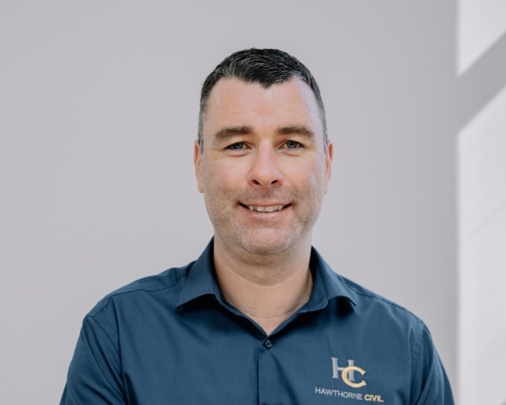 A man with short dark hair wearing a blue shirt with a Hawthorne Civil logo smiles at the camera, standing in front of a light grey background.