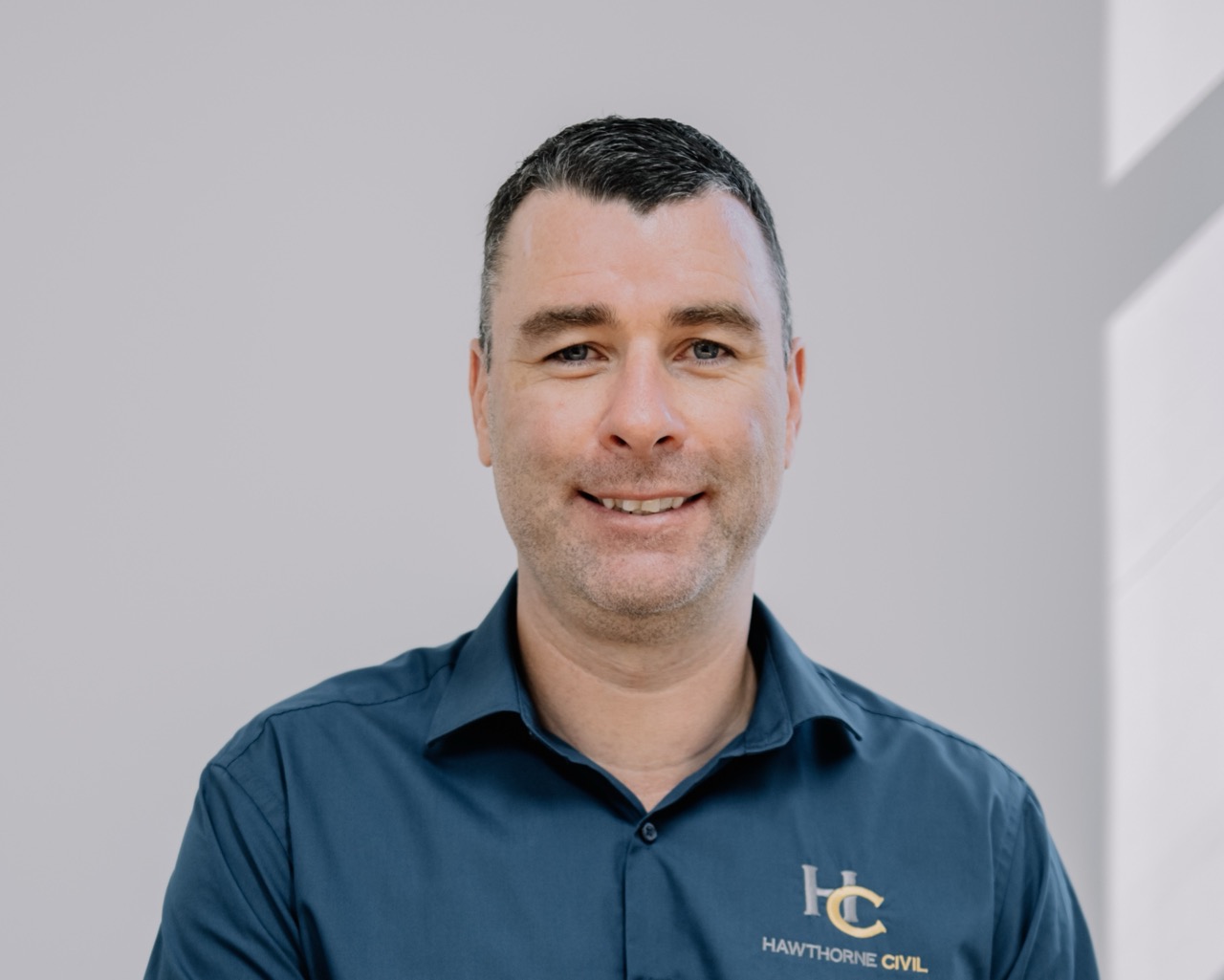 A man with short dark hair wearing a blue shirt with a Hawthorne Civil logo smiles at the camera, standing in front of a light grey background.