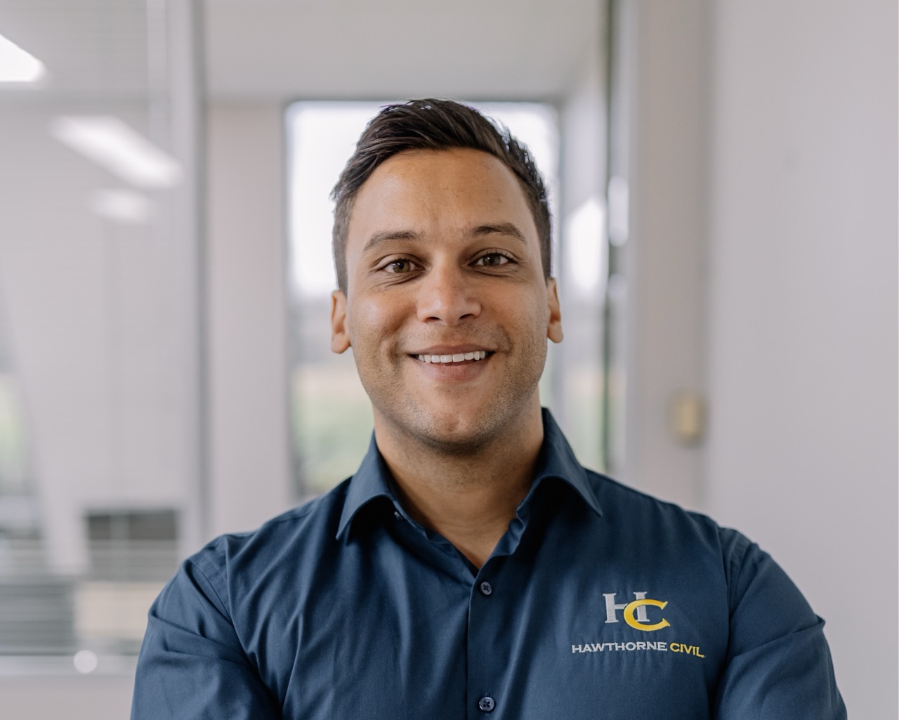 A person with short dark hair smiles confidently, standing indoors in an office. They are wearing a navy blue shirt with the Hawthorne Civil logo on the chest. The background shows windows and modern office décor.