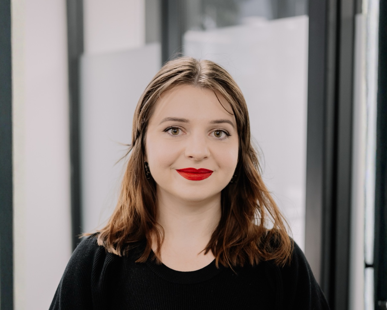 A young woman with light skin, brown hair, and red lipstick stands indoors, facing the camera and smiling softly. She is wearing a black top, and the background shows large windows with blurred light outside.