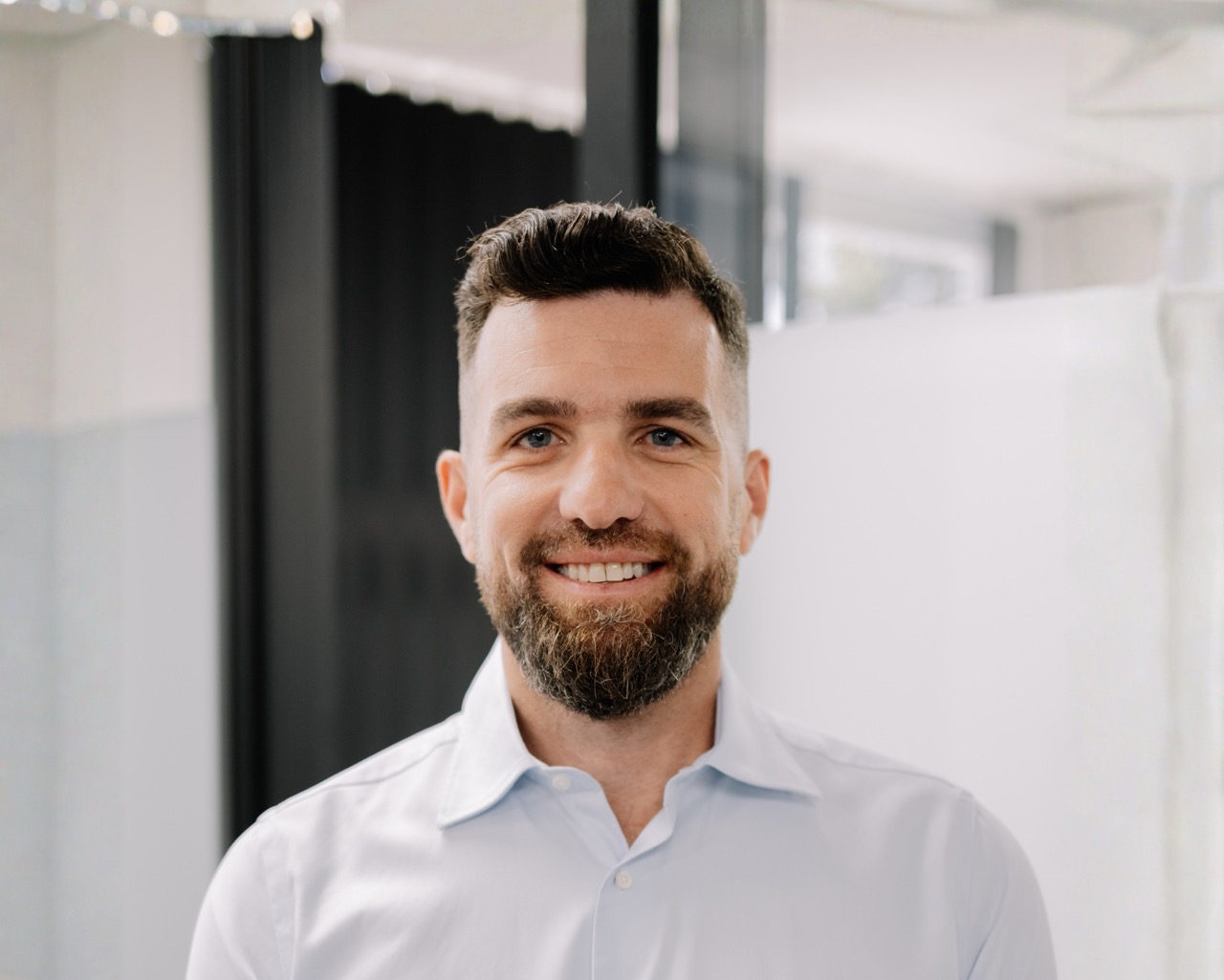 A man with short dark hair and a beard, wearing a light blue collared shirt, smiles while standing indoors in a modern, bright office environment.