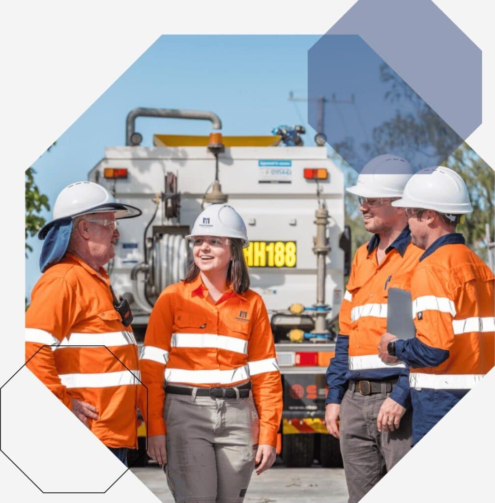 Four workers in orange high-visibility shirts and white hard hats stand outdoors, talking and smiling in front of industrial equipment and a lorry. The background features geometric shapes and a light sky.